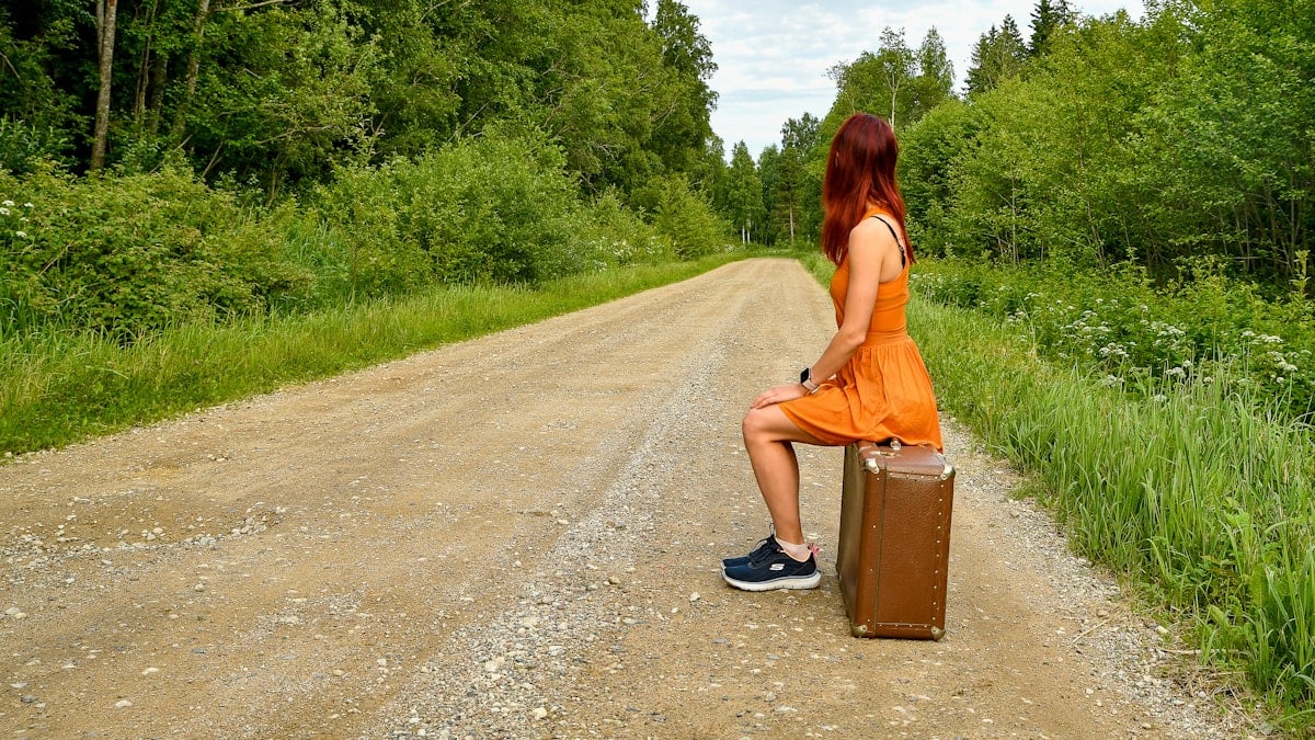 A young red-haired woman in an orange dress sitting on a vintage brown suitcase by the side of an empty gravel road, waiting during her journey. 📸 Photo by me IG: @margoevardson